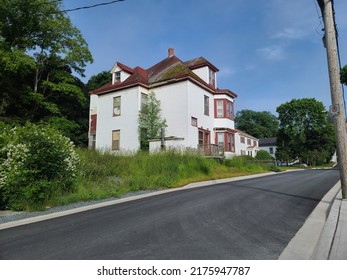 A Large Abandoned Farmhouse Home That Has Seen Better Days On A Sunny Day. The Grass Is Overgrown And The Roof Is Caving And Covered In Moss.