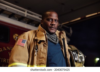 Large African American Firefighter Wearing Turnout Gear While Holding Helmet In Front Of Vintage Firetruck With Dark Background. Grim Expression On His Face At A Night Scene.