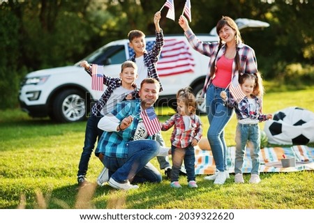 large american family spending time together. With USA flags against big suv car outdoor. America holiday. Four kids.