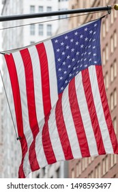 A Large American Flag Hanging Down A Pole And Waving In The Wind; In The Background, Out Of Focus Skyscrapers.