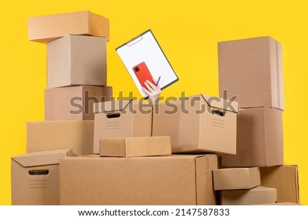 Large amount of work concept. Hand with phone and empty clipboard among boxes. Cardboard parcels of different sizes. Large number of orders. Parcels from online store. Empty clipboard on yellow.