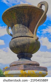 Large Ancient Iron Vase On A Concrete Plinth Stands Alone Against A Blue Cloud Sky, Looking Up From Below.