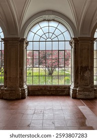 A large arched window frames a lush garden with a pink-flowering tree in the foreground. The stone columns and terracotta floor add a historic touch to the setting.
