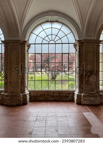 A large arched window frames a lush garden with a pink-flowering tree in the foreground. The stone columns and terracotta floor add a historic touch to the setting.