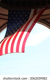 A Large Archway Leading To The Boston Harbor. There Is A Large, American Flag Hanging Down And Swaying In The Wind.