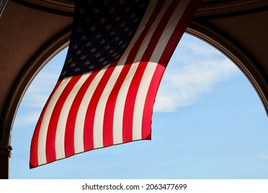 A Large Archway Leading To The Boston Harbor. There Is A Large, American Flag Hanging Down And Swaying In The Wind.