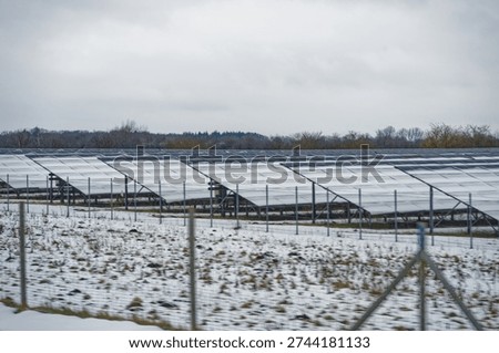 A large array of solar panels installed in a snowy field protected by a metal fence under an overcast sky during winter season.