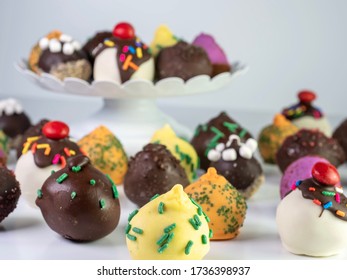 Large Assortment Of Homemade Cake Pop Balls Sitting On A Raised White Platter And Spread Out In Front Of It On A White Surface!  Delicious, Festive, Colorful Baked Goods For A Party Or Celebration!