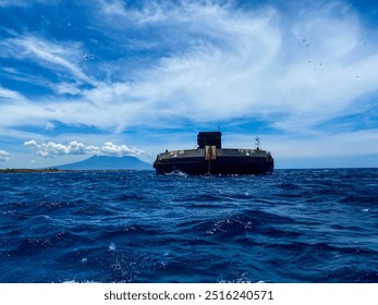 A large barge floats steadily on deep blue waters, with a distant mountain visible under a bright sky filled with wispy clouds. The ocean is slightly choppy and reflecting sunlight - Powered by Shutterstock