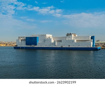 A large, barge-based correctional facility floats on calm waters under a clear blue sky, with a distant cityscape visible in the background.