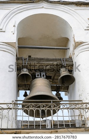 Large bell hangs within an arched stone bell tower. Smaller bells rest above, suspended from wooden beams. White pillars frame the bell chamber structure. Metal railing protects the bells below