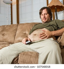 A Large Belly And A Man Smiling At It While Being Lazy With His TV Remote In Hand. Shows A Couch Potato Or A Man With A Sedentary Lifestyle .