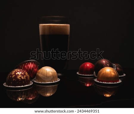 Large black coffee in a double wall glass cup with foam illuminated from above, colourful Nespresso virtue coffee pods reflecting on marble, black background. Still life and product photography.