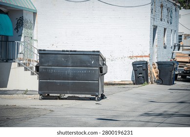 A large black dumpster and several plastic trash cans are positioned in an alleyway next to a white brick building with peeling paint