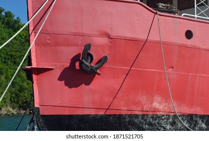 Large Black Ships Anchor In Bright Sun And Shadow Against A Bright Red Steel Hull With Rivets And White Twisted Rope Hanging Down From Cleats On The Deck. Room For Copy Space.