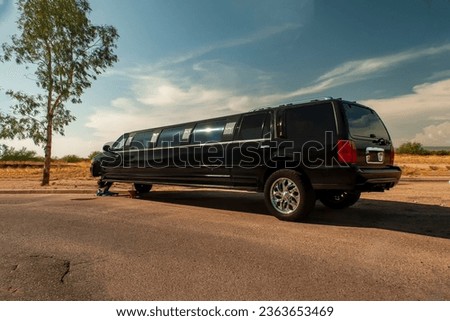 Large black stretch limousine  on a tire jack stranded on the side of a road 