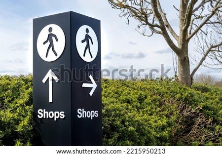 Large black and white free standing directional sign with person walking symbol and arrows pointing to shops. Outdoors in landscaped urban shopping area. 