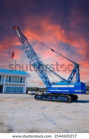 A large blue crawler crane with a long boom against a dramatic sunset sky.