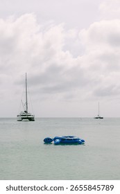 A large blue float slowly drifts in calm waters near sailboats under a cloudy sky, creating a peaceful atmosphere.