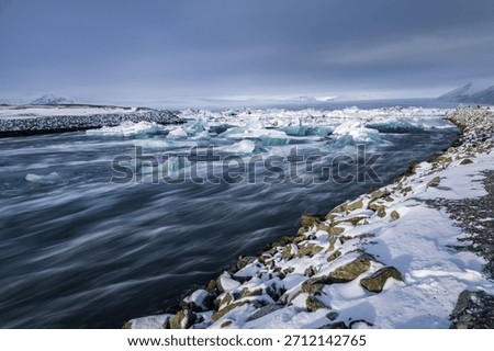 Large blue ice blocks drift through the fast-moving glacial channel at Jokulsarlon Glacier Lagoon in Iceland, surrounded by snowy shores and distant mountains during winter.