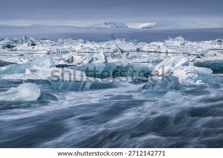 Large blue ice blocks drift through the fast-moving glacial channel at Jokulsarlon Glacier Lagoon in Iceland, surrounded by snowy shores and distant mountains during winter.