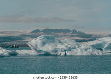 Large blue icebergs float serenely in a glacial lagoon under a cloudy sky. Backdrop of Icelandic mountains. The scene highlights the cold, isolated beauty of this icy landscape - Powered by Shutterstock