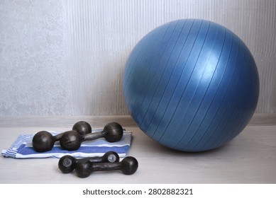 Large blue stability exercise ball with set of black dumbbells and folded towel on light wooden floor against neutral wall. Home fitness and workout equipment setup.