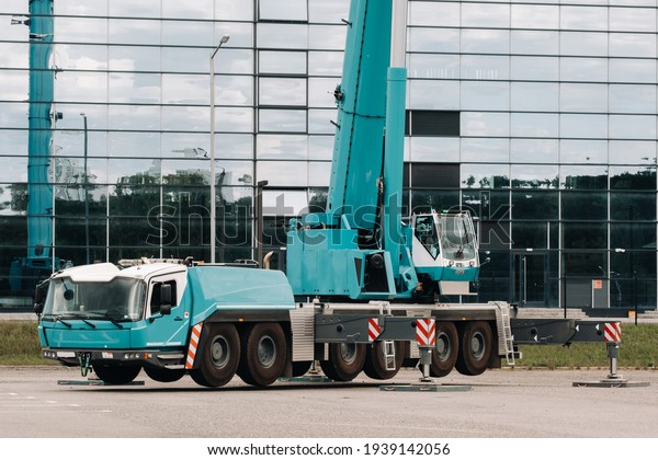 A large blue truck\
crane stands ready to operate on hydraulic supports on a platform\
next to a large modern building. The largest truck crane for\
solving complex tasks