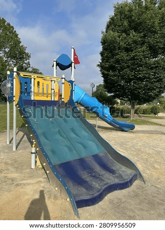 A large blue wave-textured slide with a cylindrical blue tube slide beside it is the centerpiece of a colorful playground set featuring yellow and blue panels. The equipment stands on a sandy ground under a clear blue sky, with lush green trees and a clock pole visible in the background of the suburban park.