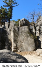 A large boulder rests on a flat rock surface next to a stone staircase and trees.