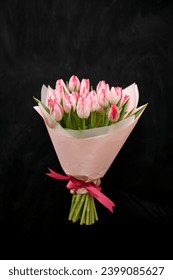 Large bouquet of pink tulips in a pink wrapper is held in front of a female hand dressed in black clothes on a black background