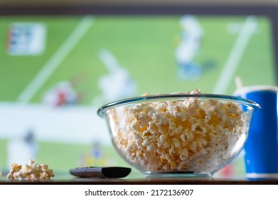 A Large Bowl Of Popcorn And A TV Remote Against The Background Of A Large TV Screen With A Sports Match. Watching Sports Programs On TV - Football, Basketball, American Football. Fast Food, Snacks.