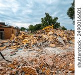 A large brick building has just been demolished. A pile of red bricks, wood insulation and debris are stacked in a pile. Bulldozer in background of view. Large demolition construction site.