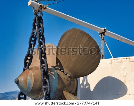 A large bronze ship propeller hangs suspended by heavy iron chains against a vibrant blue sky. The metallic blades of the industrial maritime component show weathered textures under direct sunlight on a deck.