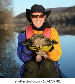 A large brown flathead catfish being held horizontally facing the viewer by a smiling woman sitting in a canoe in a blue and gold dry suit on a mirrored river in winter