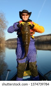 A large brown flathead catfish being held vertically by a smiling woman standing in a canoe in a blue and gold dry suit on a mirrored river in winter
