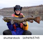 A large brown olive colored flathead catfish fish being held horizontally by a smiling woman on a smooth river