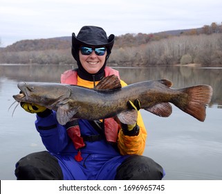 A large brown olive colored flathead catfish fish being held horizontally by a smiling woman on a smooth river