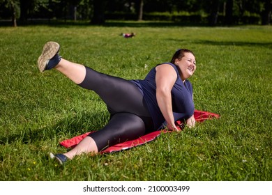 Large Build Woman Is Making Yoga Exercises On Mat On Grass In Public Park And Smiling.