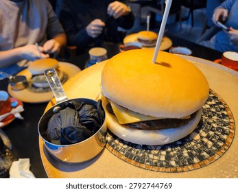 Large burger sits on patterned plate. Small pot holds black gloves beside it. People dine in background, blurred. Wooden table holds meal setup. Burger has toothpick through top