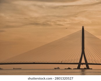 A large cable-stayed bridge stands silhouetted against a vibrant orange sunset sky over a calm body of water with distant ships. The modern suspension architecture is highlighted by the warm golden hour light.
