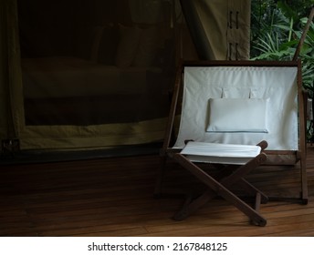 Large camping wooden chair with white fabric seat and footrest on the terrace in front of the room of large tent. Folding outdoor sunbed on white glamping tent with copy space.