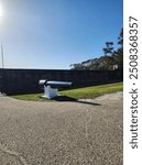 A large cannon mounted on a white base, pointing towards a brick fortification wall under a clear blue sky. The area is paved with gravel and a grassy patch is visible.