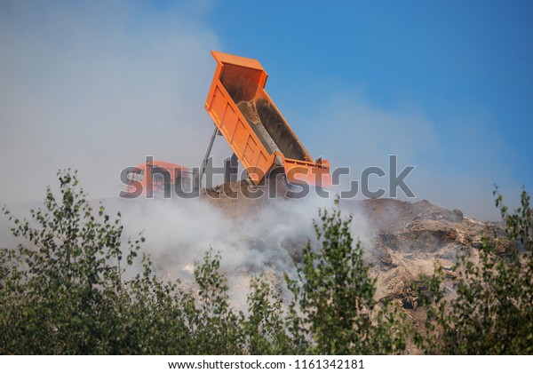 a
large car dump truck on a sandy quarry, during a fire at a landfill
helps to fight fire, dropping on the flames from the raised body of
a large pile of land and sand, all in smoke and
dust
