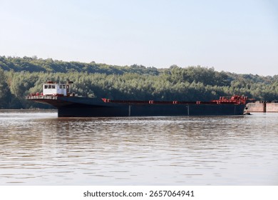 A large cargo ferry navigating calm waters of a river with a forested background, captured on a clear sunny day, highlighting transportation, nature, and serene environment