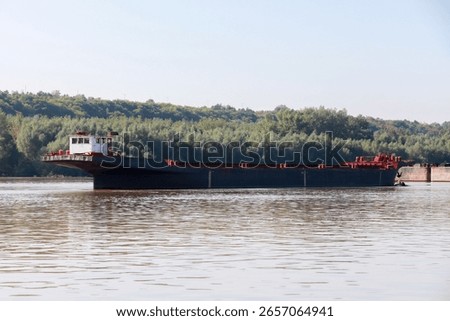 A large cargo ferry navigating calm waters of a river with a forested background, captured on a clear sunny day, highlighting transportation, nature, and serene environment
