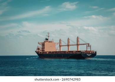 A large cargo ship with multiple cranes sails across a vast expanse of blue ocean water under a partly cloudy sky.