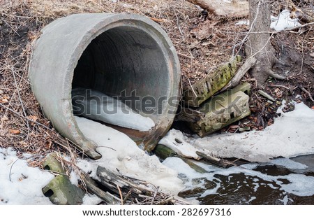 Large cement sewer drain pipe emptying into foamy puddle with debris in winter, surrounded by branches, dirt, and patches of snow.