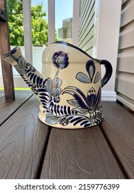 A Large, Ceramic, Watering Can That Is Sitting On A Composite Wood Deck In A Suburban Backyard.