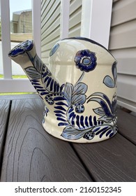 A Large, Ceramic, Watering Can That Is Sitting On A Composite Wood Deck In A Suburban Backyard.